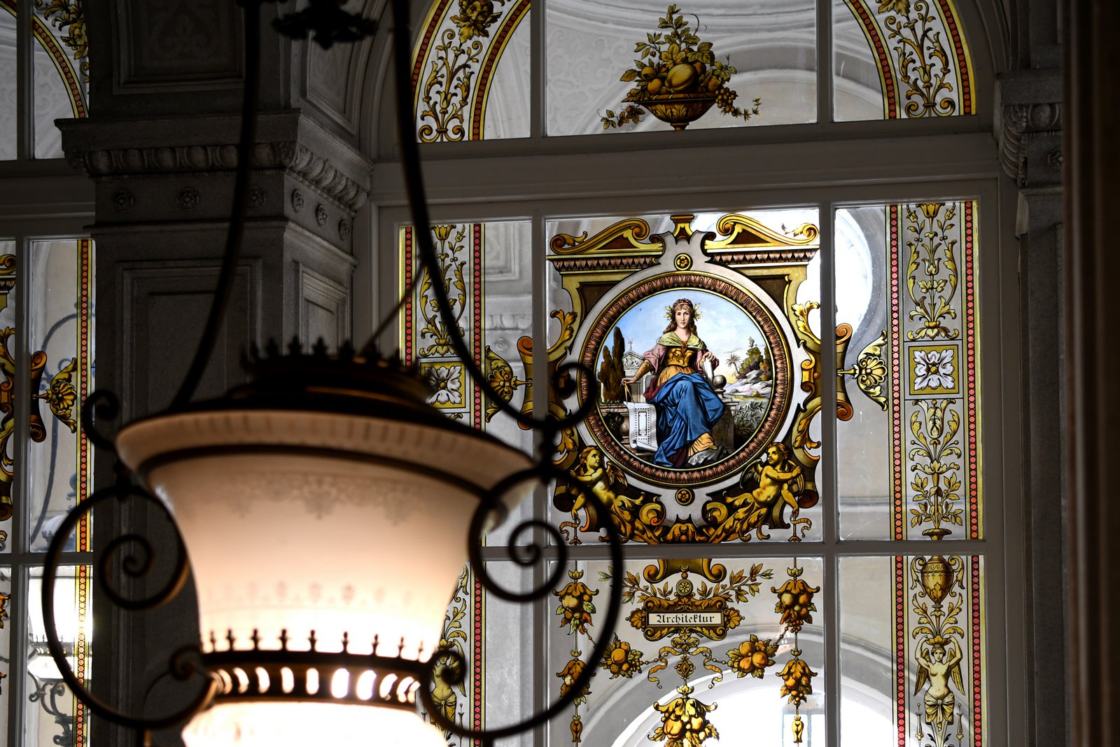 Zbliżenie na witraż – centralna postać w medalionie, dekoracyjne bordiury roślinne; na pierwszym planie rozmyty, ozdobny klosz lampy.