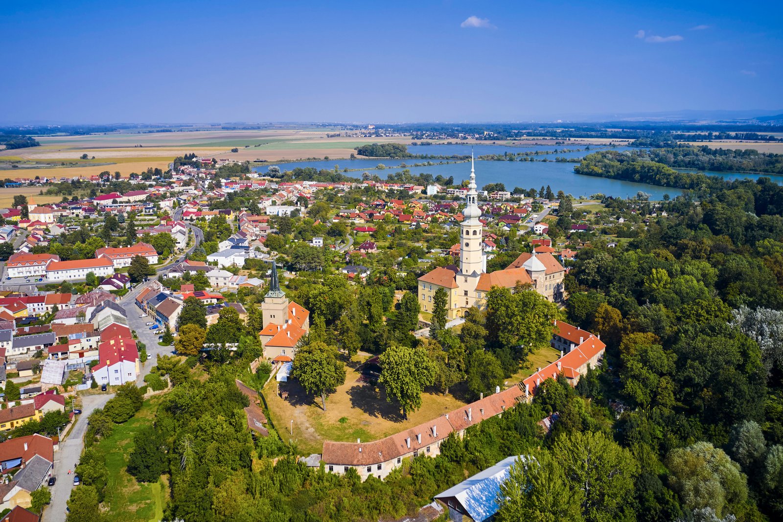 Ujęcie z drona – panorama miasteczka z zamkiem i wysoką wieżą pośrodku oraz dużym jeziorem/stawami w tle; wokół zieleń i zabudowa Haná.