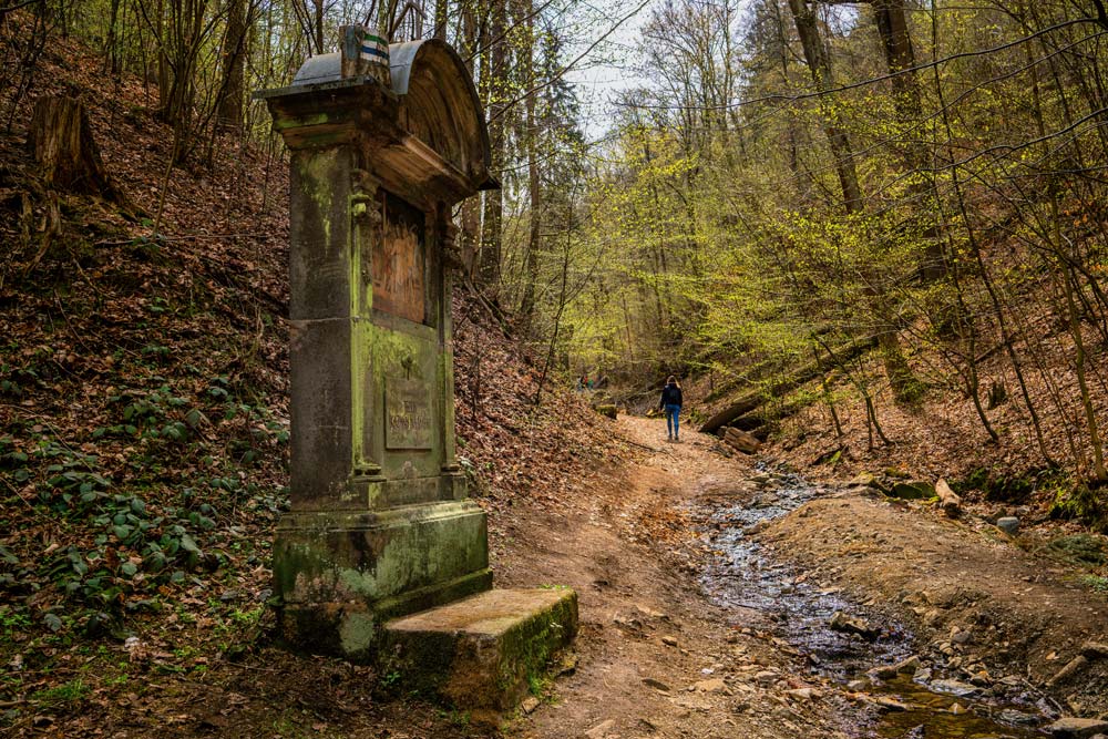 Leśny jar z potokiem i ścieżką; na pierwszym planie kamienna kapliczka Drogi Krzyżowej, w oddali wędrowcy.