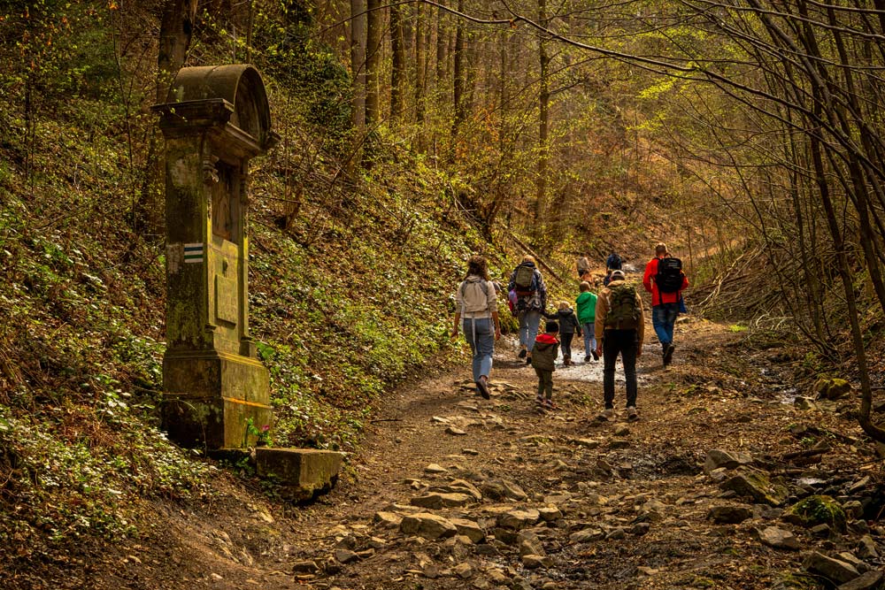 Grupa turystów idzie leśną ścieżką w wąwozie; po lewej kamienna kapliczka stacji Drogi Krzyżowej.