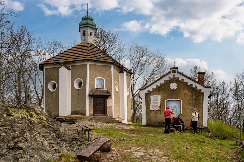 Widok na kaplicę na Bardzkiej Kalwarii oraz sąsiedni budynek z tablicą informacyjną; turyści odpoczywają na ławkach.