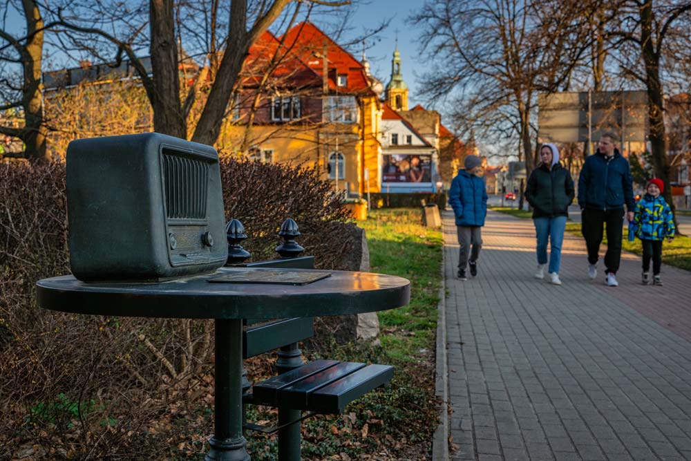 Mosiężne radio na okrągłym stoliku obok ławki; w tle rodzina spaceruje alejką w Dzierżoniowie.