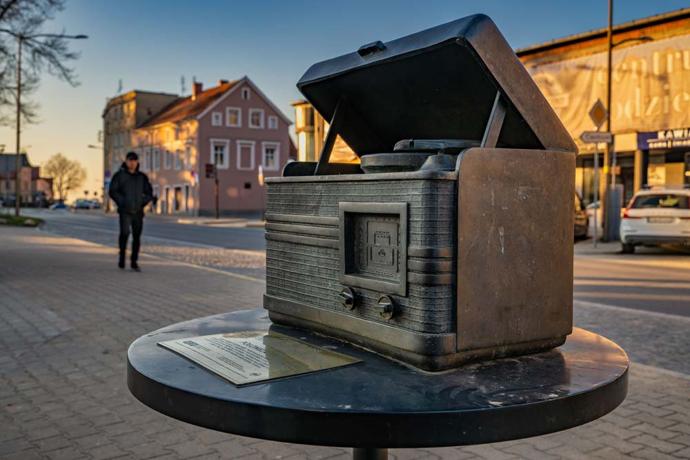 Mosiężna rzeźba retro radioodbiornika z podniesioną klapą na okrągłym cokole; w tle ulica i zabudowa Dzierżoniowa.
