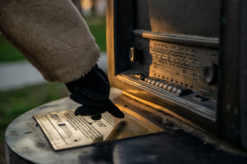 Zbliżenie na mosiężną tabliczkę informacyjną Traktu Diora; dłoń w rękawiczce dotyka opisu przy rzeźbie radia.