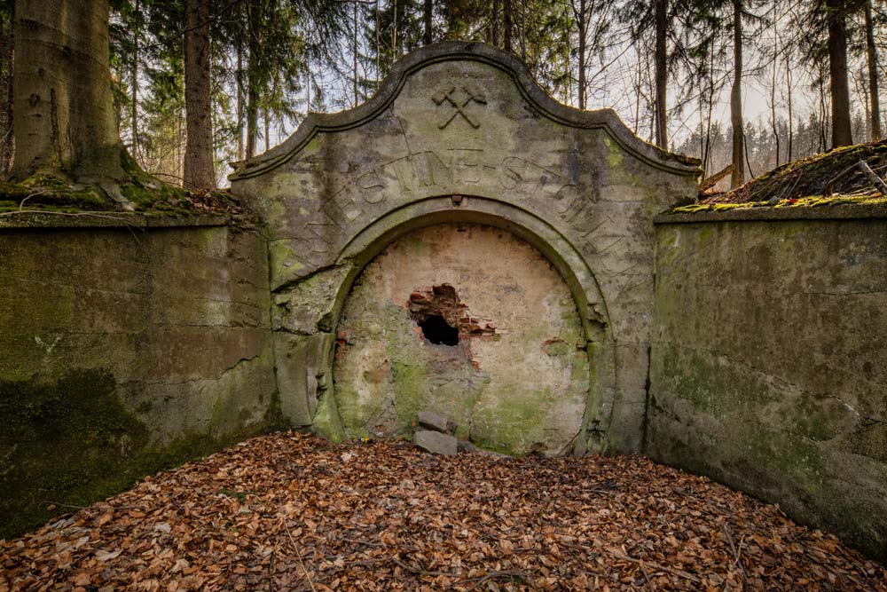 Frontowy widok na portal Ernestine-Stollen: łuk z napisem, zamurowane wejście z ubytkiem i leśne tło.