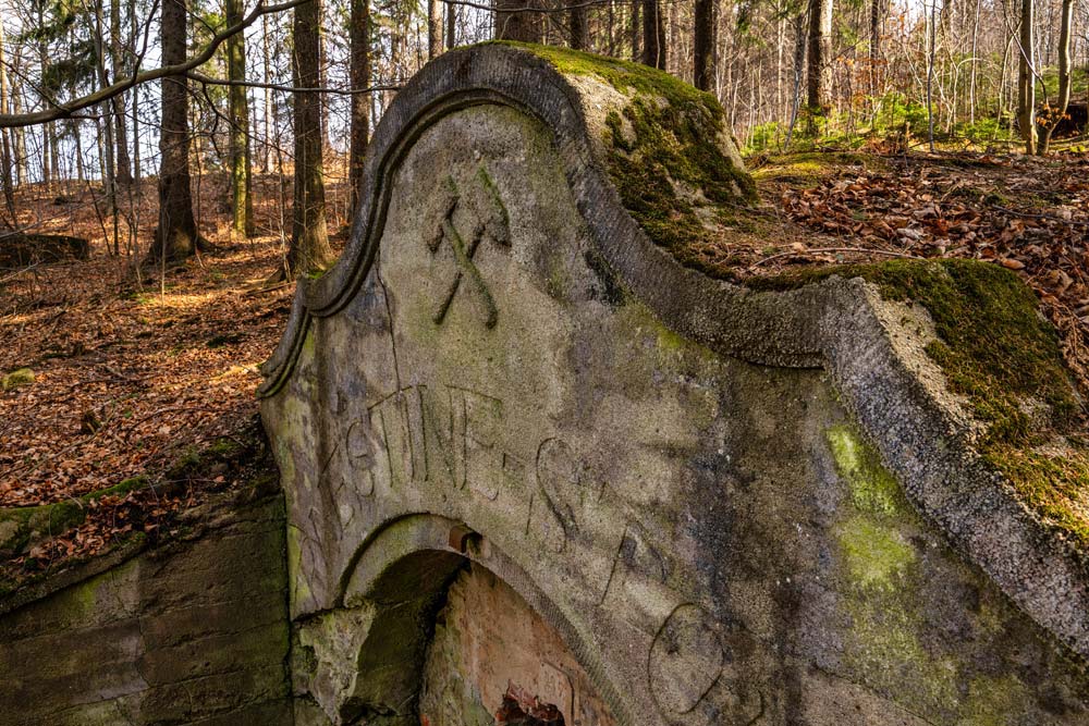 Zbliżenie na górną część portalu Ernestine-Stollen z wyrytym napisem i symbolem górniczym, porośniętą mchem.