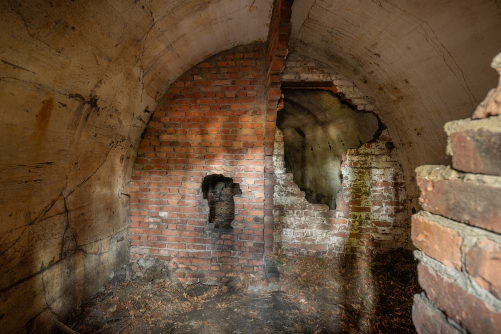 Wnętrze zniszczonego korytarza sztolni: sklepienie z betonu, ceglana ściana i przebite otwory w murze.