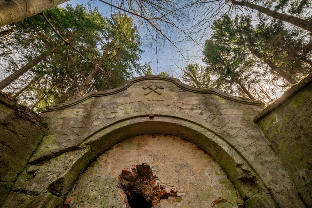 Kamienny portal „Ernestine-Stollen” widziany z dołu; nad łukiem skrzyżowane młotki, wokół korony drzew.