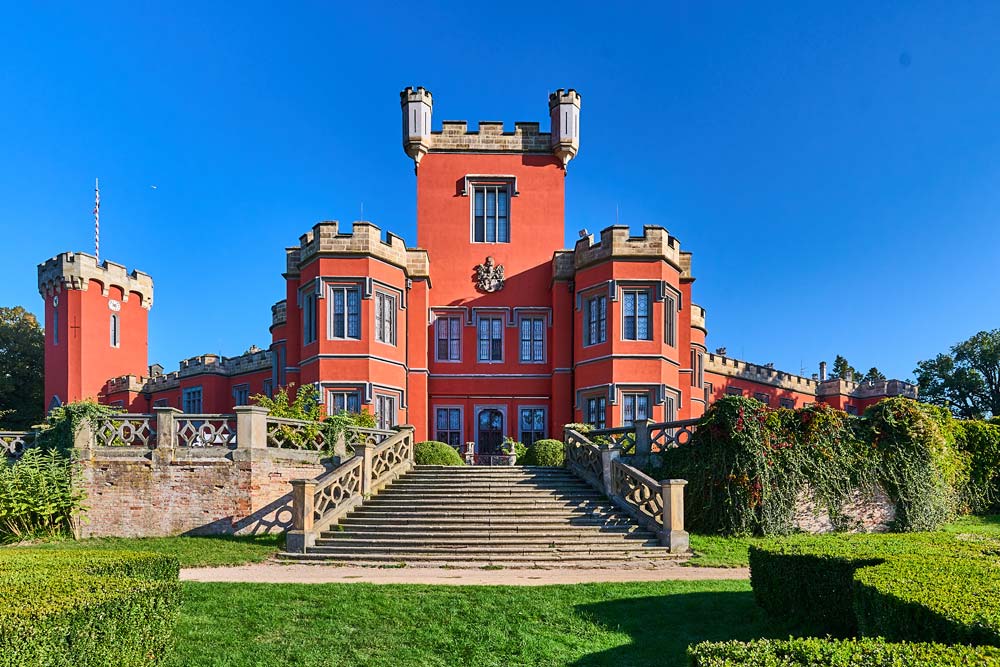 Wejście do zamku Hradec u Nechanic: szerokie schody prowadzą do czerwonej fasady z wieżą i blankami.