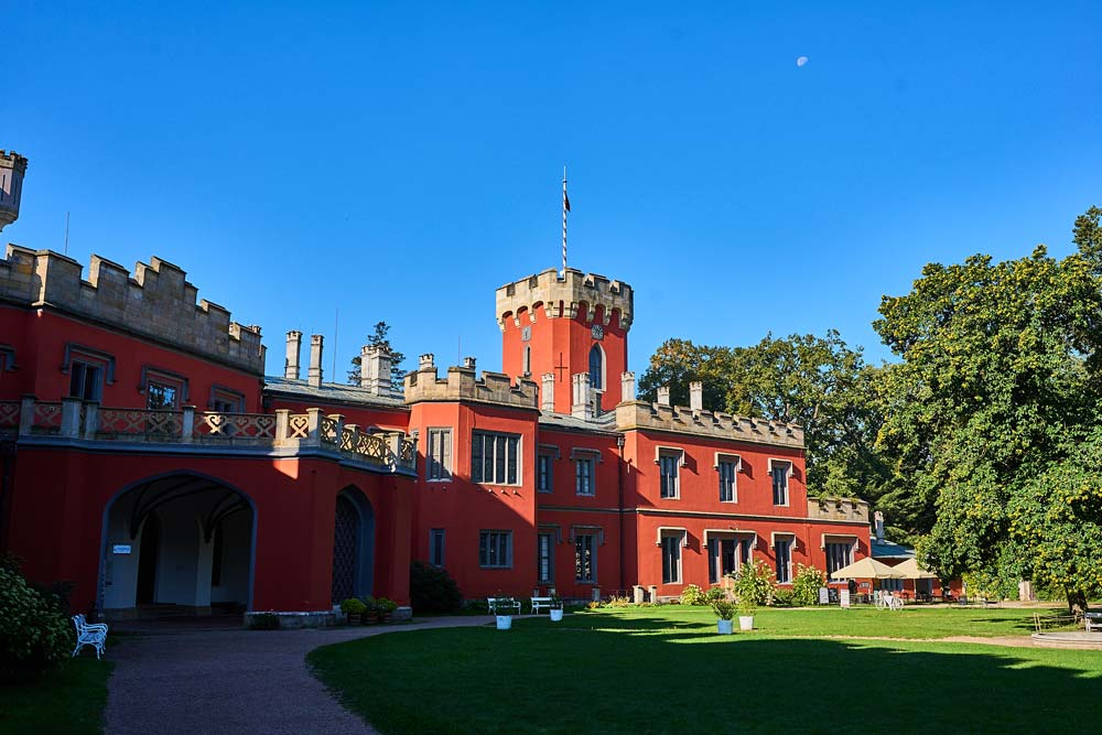 Czerwona, neogotycka fasada zamku Hradec u Nechanic z wieżą i blankami, na tle parku i błękitnego nieba.