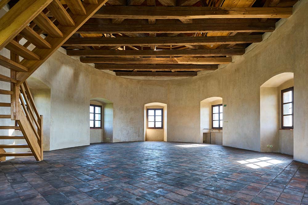 Pusta sala na poddaszu zamku Humprecht: drewniane belki stropu, schody i rząd okien nad kamienną posadzką.