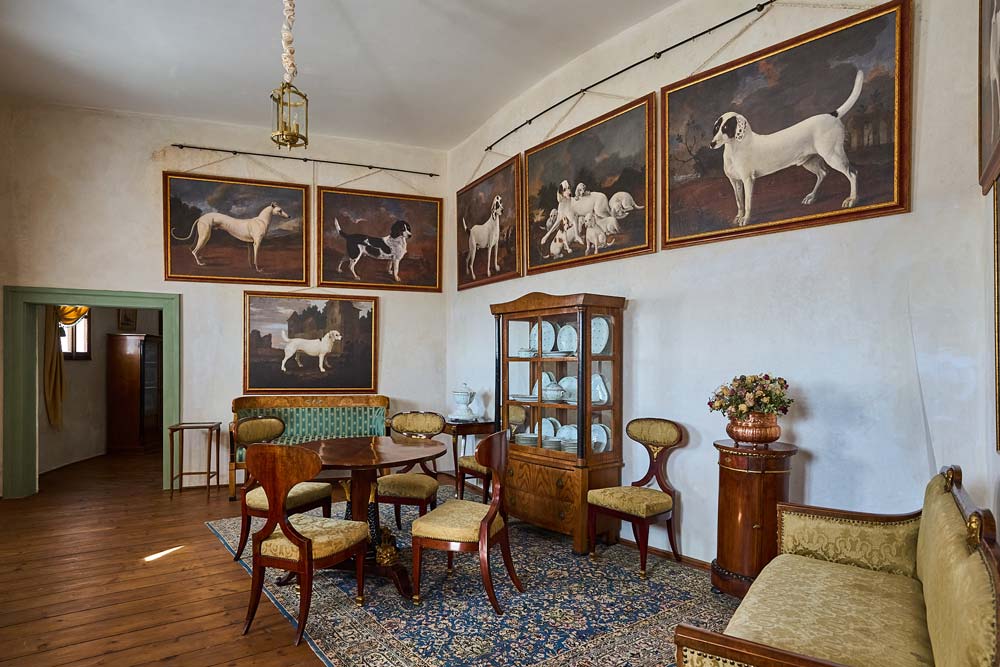 Salon w Humprechcie z obrazami psów myśliwskich na ścianach, okrągłym stołem i witryną z porcelaną.