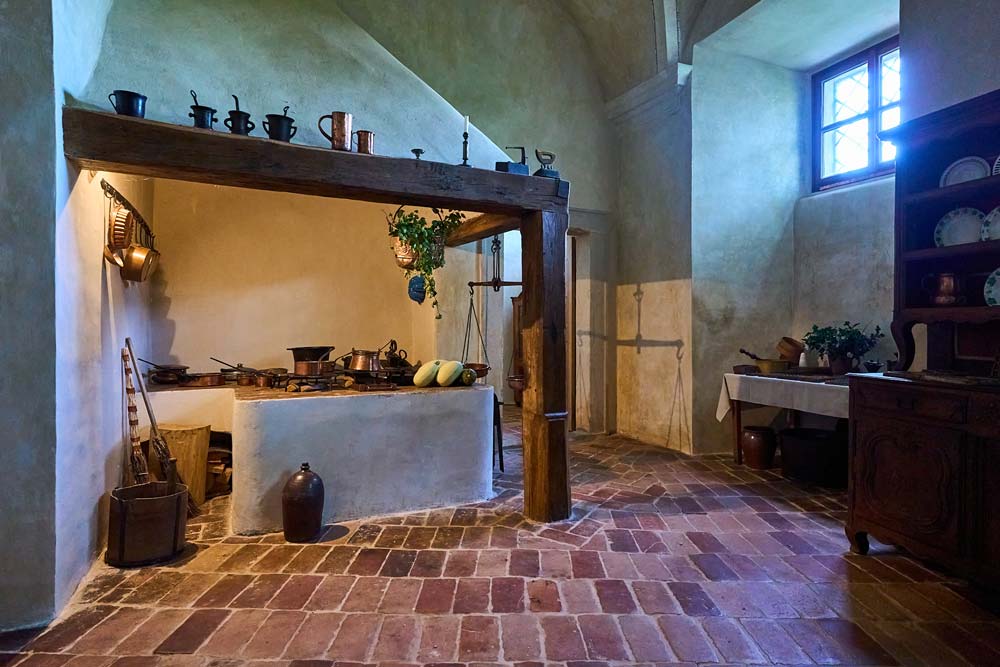 Czarna kuchnia w zamku Humprecht: murowane palenisko pod drewnianą belką, miedziane garnki i ceglana posadzka.