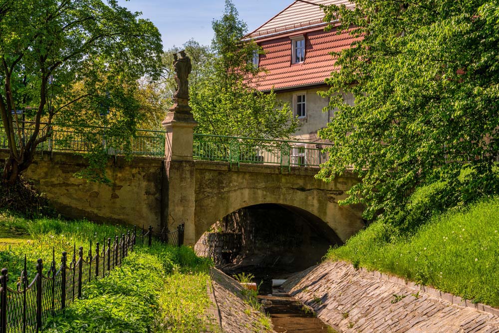 Perspektywa na most nad potokiem Zatoka: kamienna arkada, zielone zbocza i figura Matki Bożej na przyczółku.