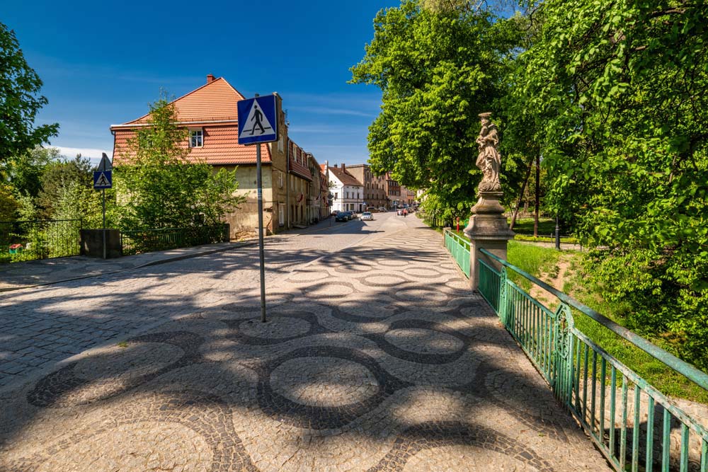 Widok ulicy Ziębickiej z ozdobną kostką i znakiem przejścia; po prawej zielona balustrada i figura na moście.