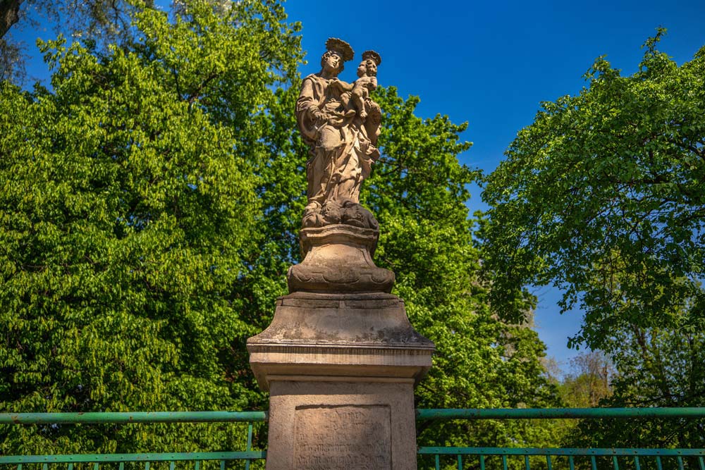 Zbliżenie na rzeźbę Matki Bożej z Dzieciątkiem na Moście Ziębickim na tle błękitnego nieba i koron drzew.