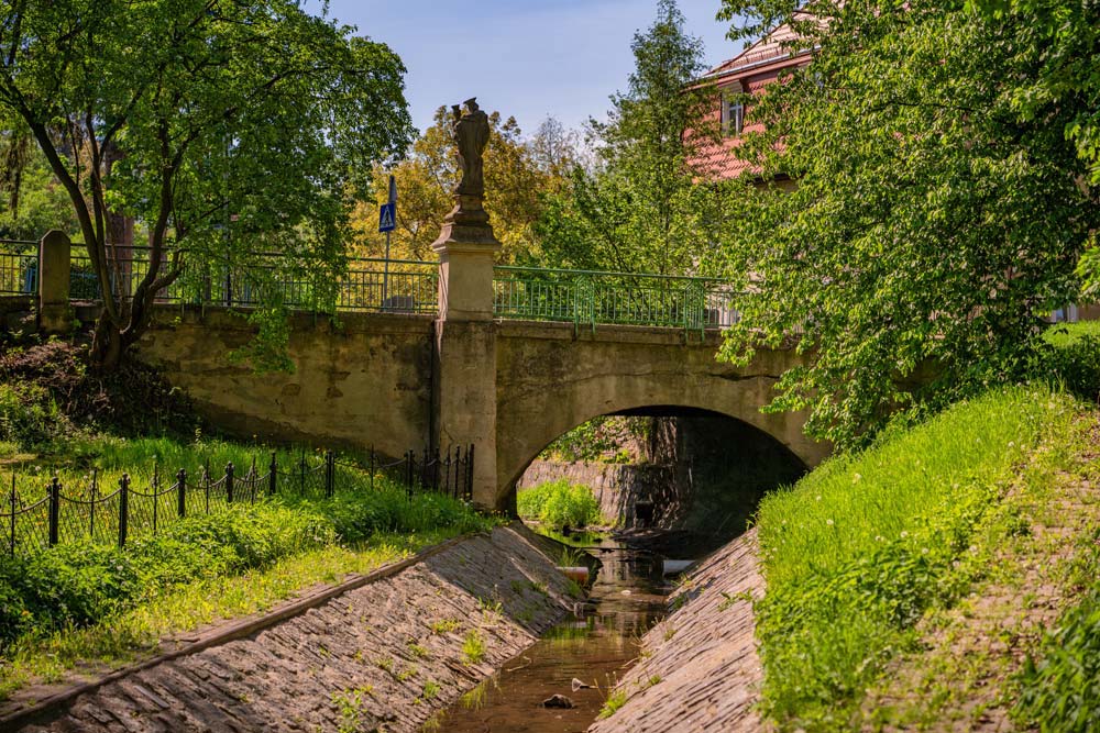 Kamienny most o łukowej arkadzie nad potokiem Zatoka; na skraju przeprawy stoi figura Maryi, wokół zielone skarpy.