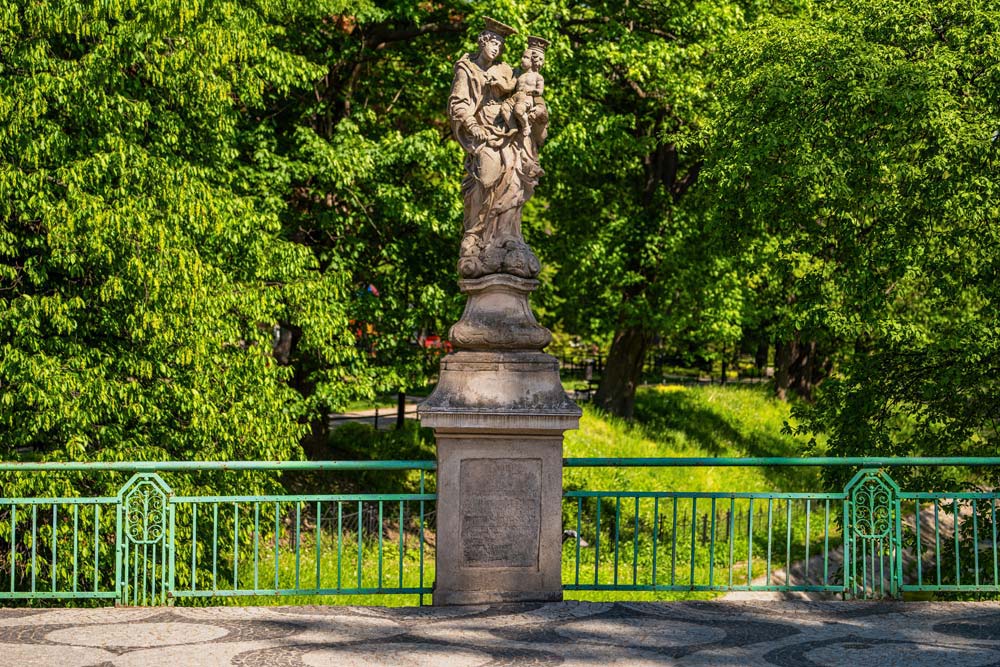 Barokowa figura Matki Bożej z Dzieciątkiem na cokole przy zielonej balustradzie Mostu Ziębickiego, wśród bujnych drzew.