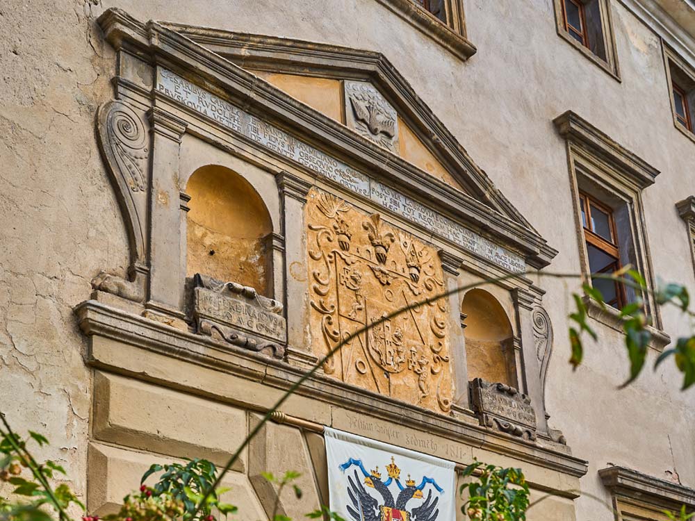 Renesansowy portal z herbem na fasadzie Staré Hrady; pod nim wisi baner, a w tle widoczne okna pałacu.