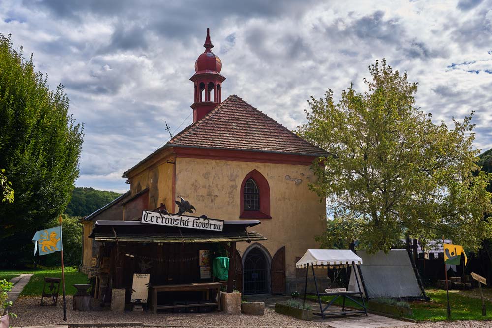 „Čertovská kovárna” przy zamku Staré Hrady: mały budynek z wieżyczką, szyldem i namiotami w plenerze.