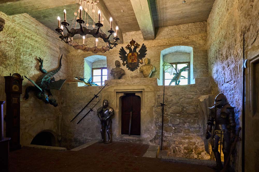 Sala zamkowa z surowymi kamiennymi murami: zbroje rycerskie przy wejściu, miecze na ścianach i dekoracyjny smok pod żyrandolem.