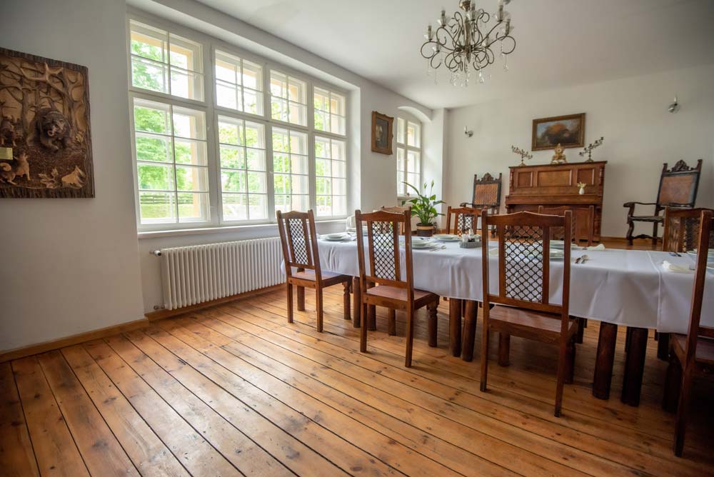 Jadalnia w Zamku Bożejów z długim stołem nakrytym białym obrusem, drewnianymi krzesłami i dużymi oknami.