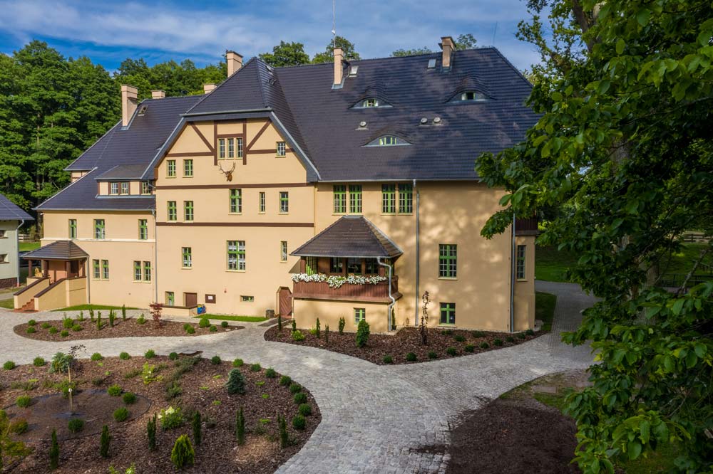 Boczne skrzydło Zamku Bożejów z balkonem i kwiatami; brukowana alejka prowadzi przez świeżo nasadzone rabaty.