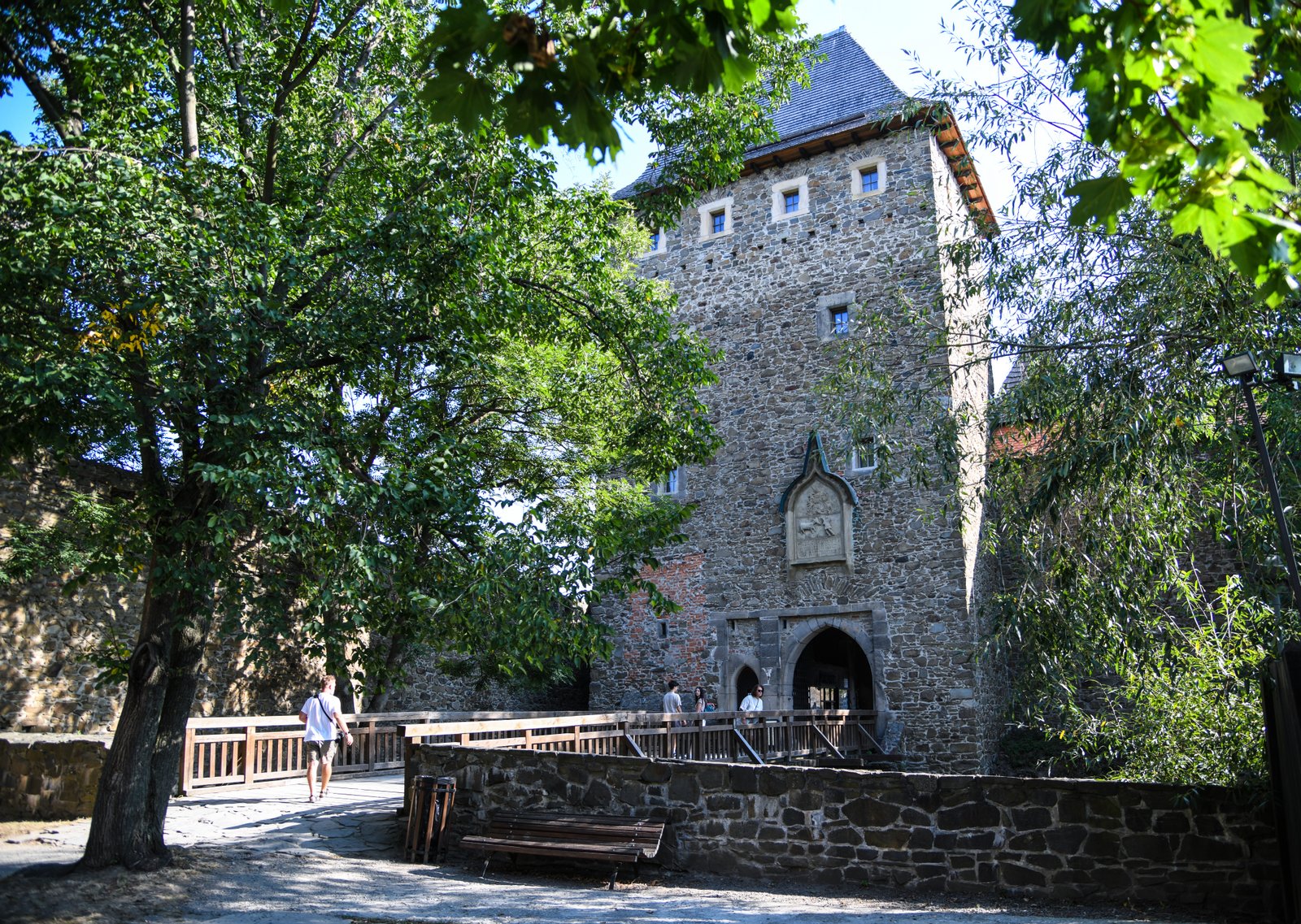 Wejście do zamku Helfštýn: kamienna brama z wysoką wieżą i drewnianym mostem w cieniu drzew.