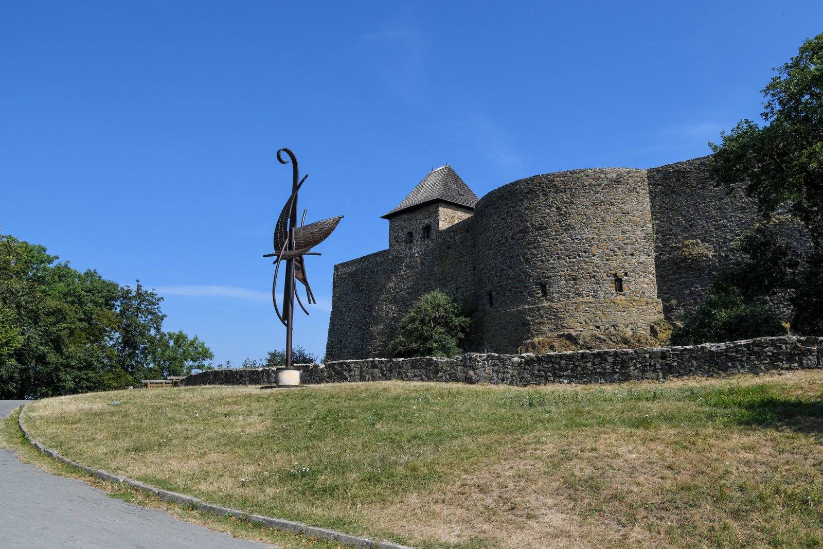 Fragment murów obronnych zamku Helfštýn i nowoczesna metalowa rzeźba na trawiastym zboczu pod błękitnym niebem.