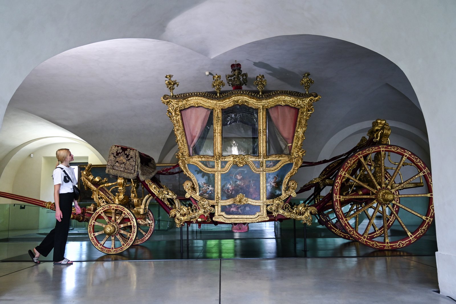 Złota barokowa kareta biskupia w sali muzealnej; zwiedzająca stoi obok bogato zdobionego pojazdu z wielkimi kołami.