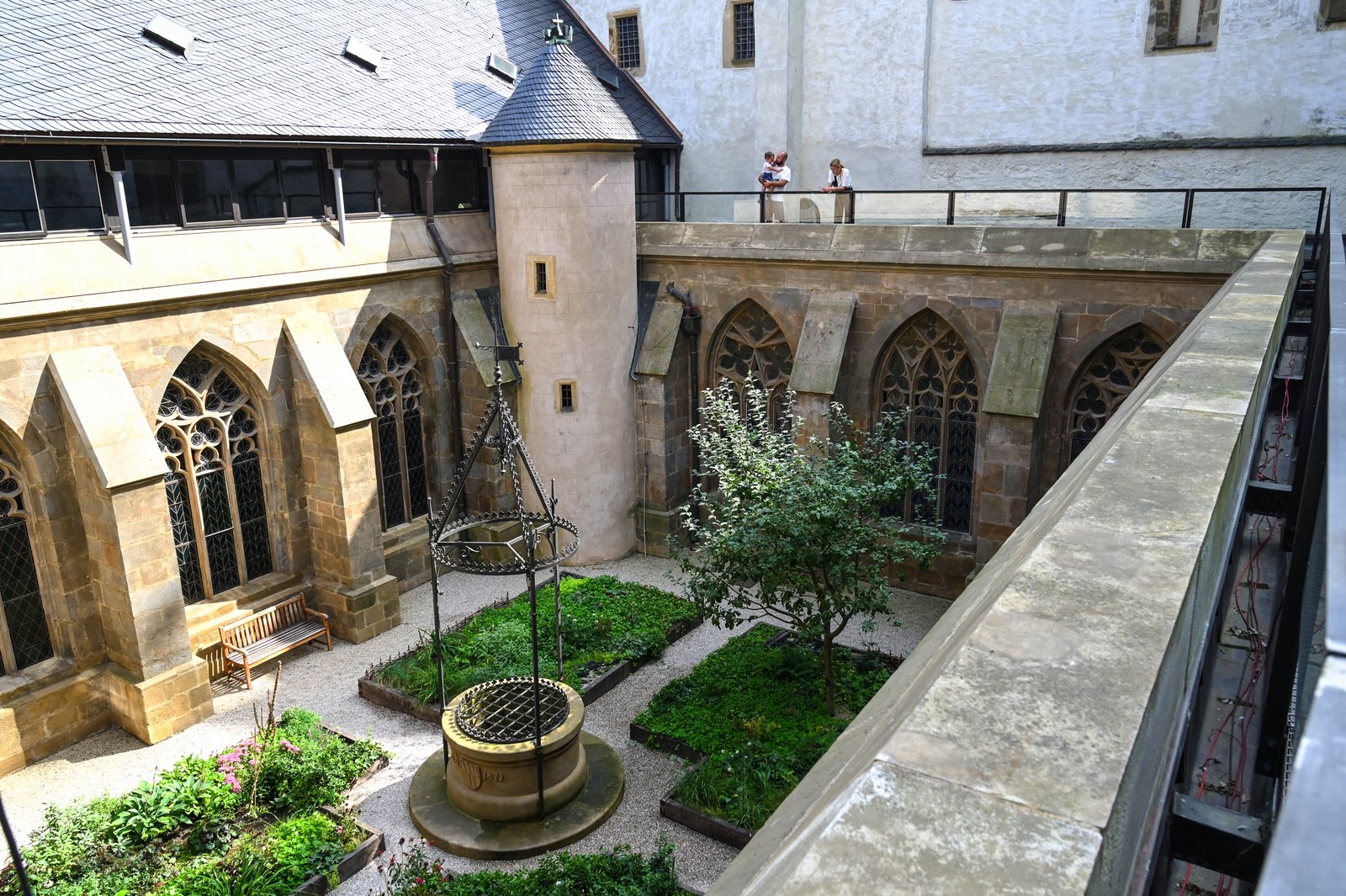 Dziedziniec rajski widziany z góry: studnia, rabaty i drzewo, wokół gotyckie okna; na galerii stoją zwiedzający.