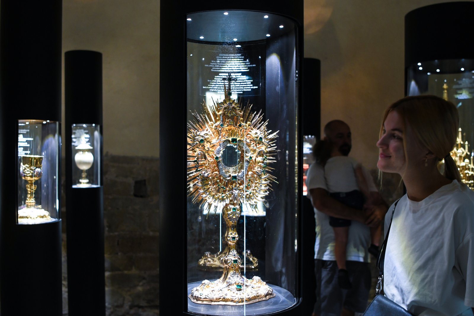 Skarbiec muzeum: bogato zdobiona monstrancja w podświetlanej gablocie, obok zwiedzająca; w tle inne kielichy liturgiczne.