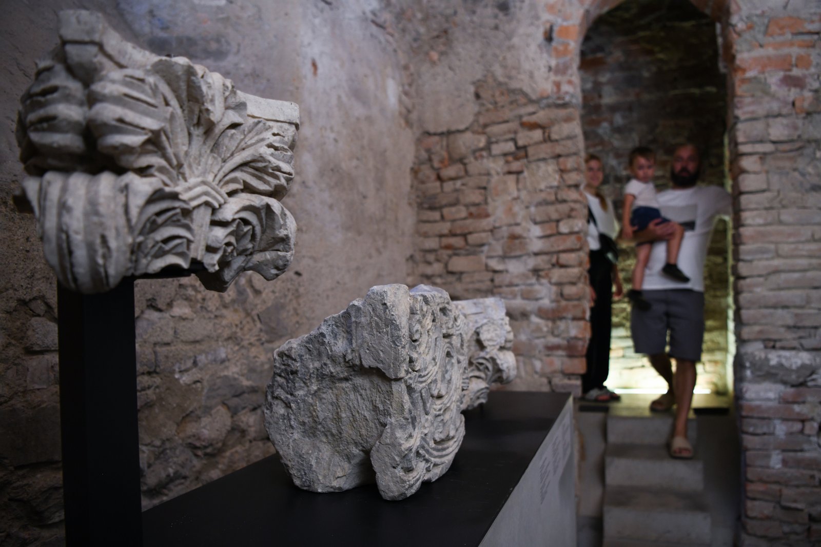 Podziemne przejście z cegły i kamienia; na postumentach leżą fragmenty rzeźb, a w tle rodzina schodzi po schodach.