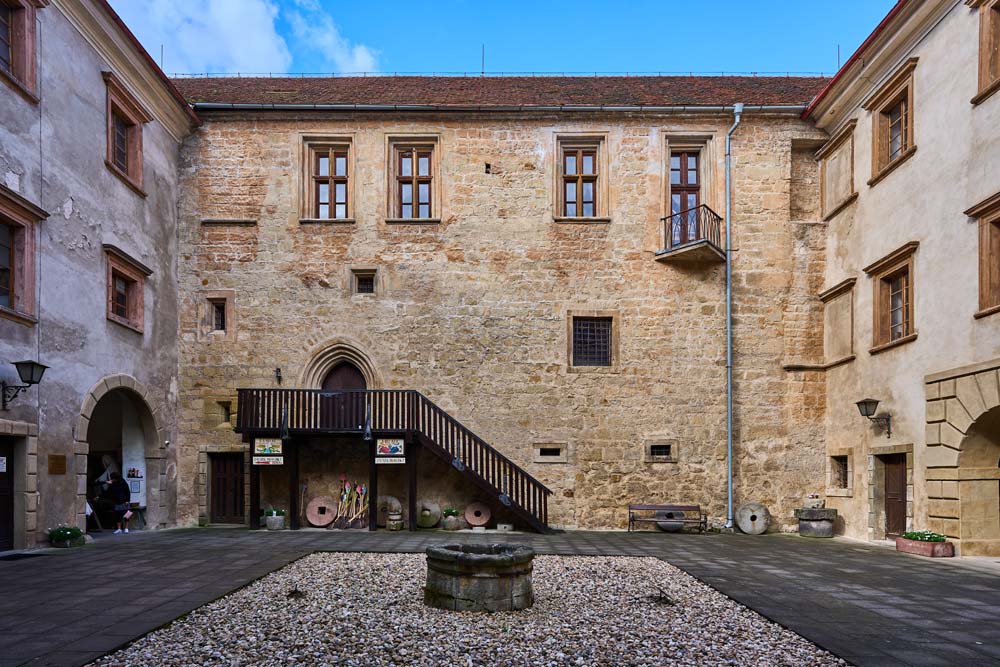 Wewnętrzny dziedziniec Staré Hrady z kamiennym skrzydłem i drewnianymi schodami; na środku niska studnia w żwirowym otoku.