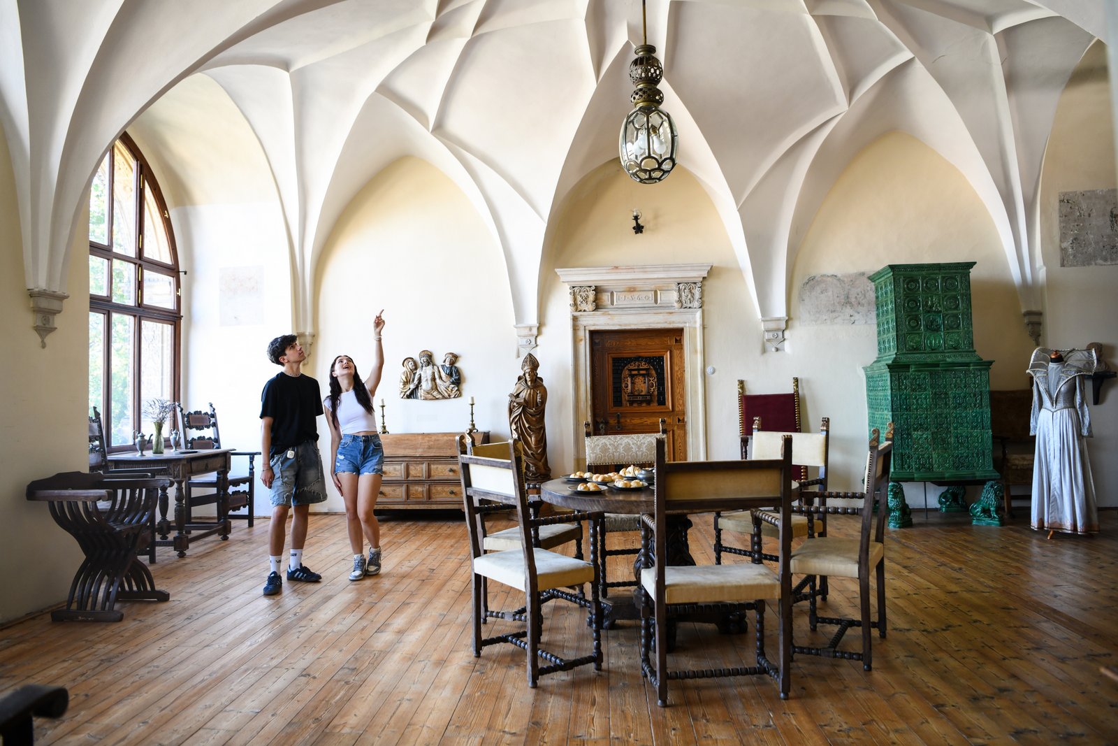 Reprezentacyjna sala Zamku Šternberk ze sklepieniem krzyżowym, stołem i antycznymi meblami; para zwiedzających wskazuje detale.