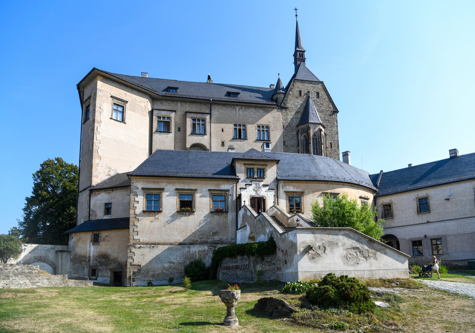 Widok na zabudowania Zamku Šternberk: schody na taras, masywne mury i wieża na tle błękitnego nieba.