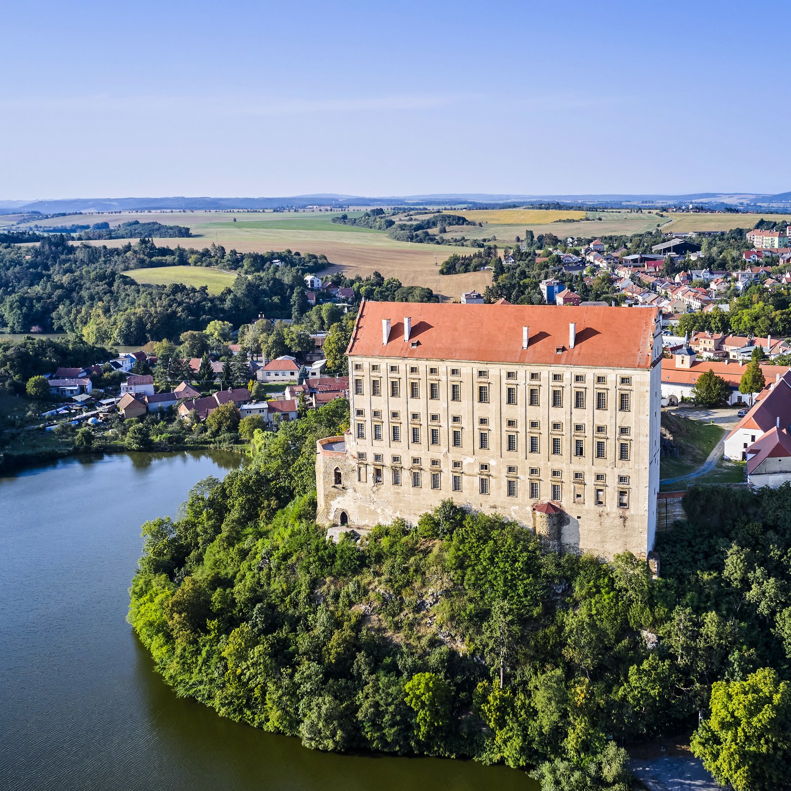 Zdjęcie z drona pokazujące Pałac Plumlov górujący nad jeziorem i miasteczkiem na Morawach; smukła bryła zamku wyraźnie dominuje w krajobrazie.
