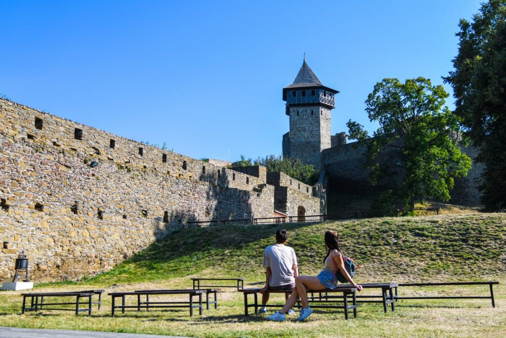 Dziedziniec Helfštýna i potężne mury obronne; para turystów siedzi na ławkach, a w tle widać wieżę.