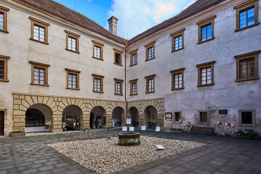 Dziedziniec zamku Staré Hrady: jasne elewacje z rzędami okien, arkadowe podcienia i kamienna studnia pośrodku.