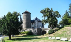 Cylindryczna wieża i mury Zamku Šternberk widziane z parku; na trawie stoją białe ławki i rosną wysokie drzewa.