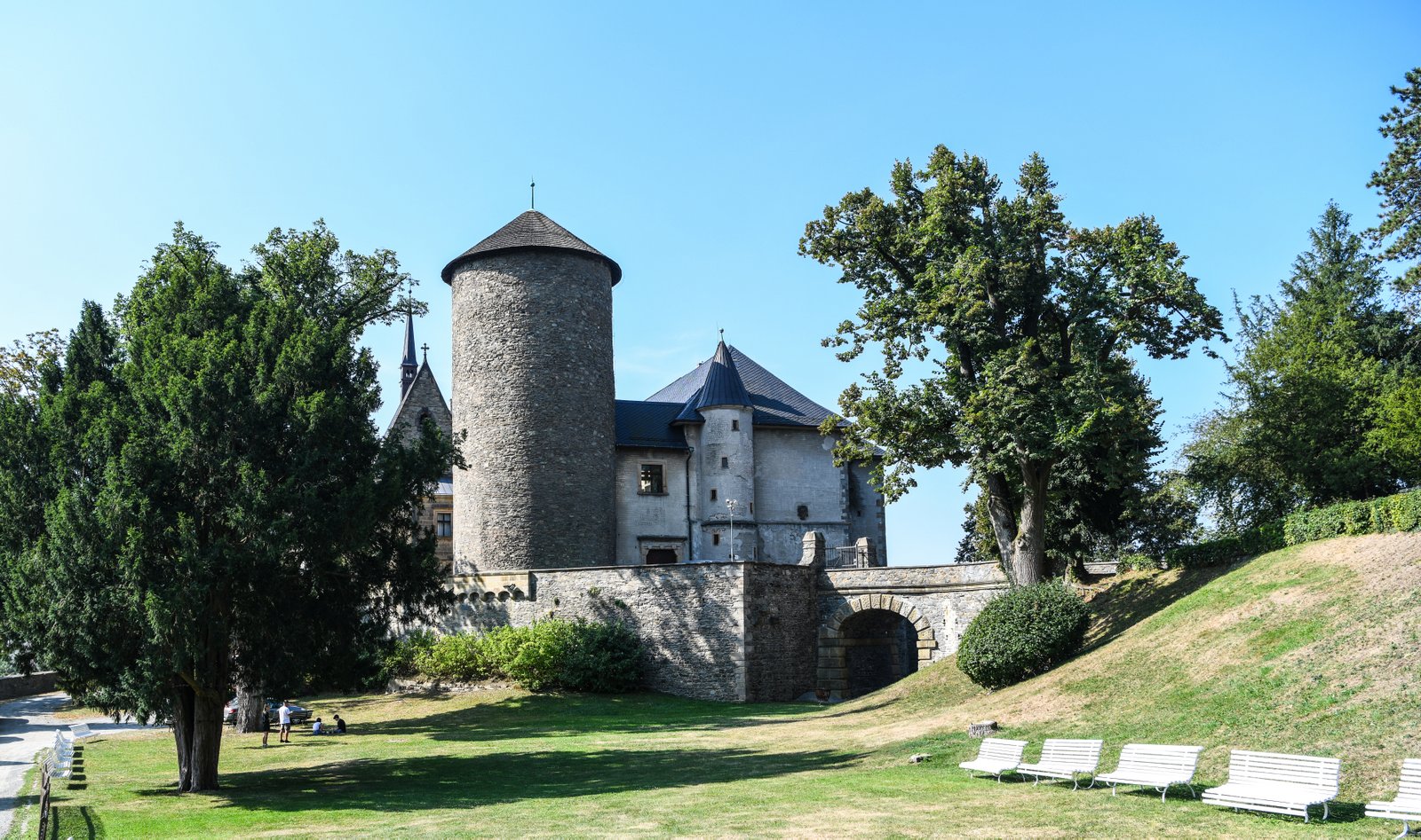 Cylindryczna wieża i mury Zamku Šternberk widziane z parku; na trawie stoją białe ławki i rosną wysokie drzewa.