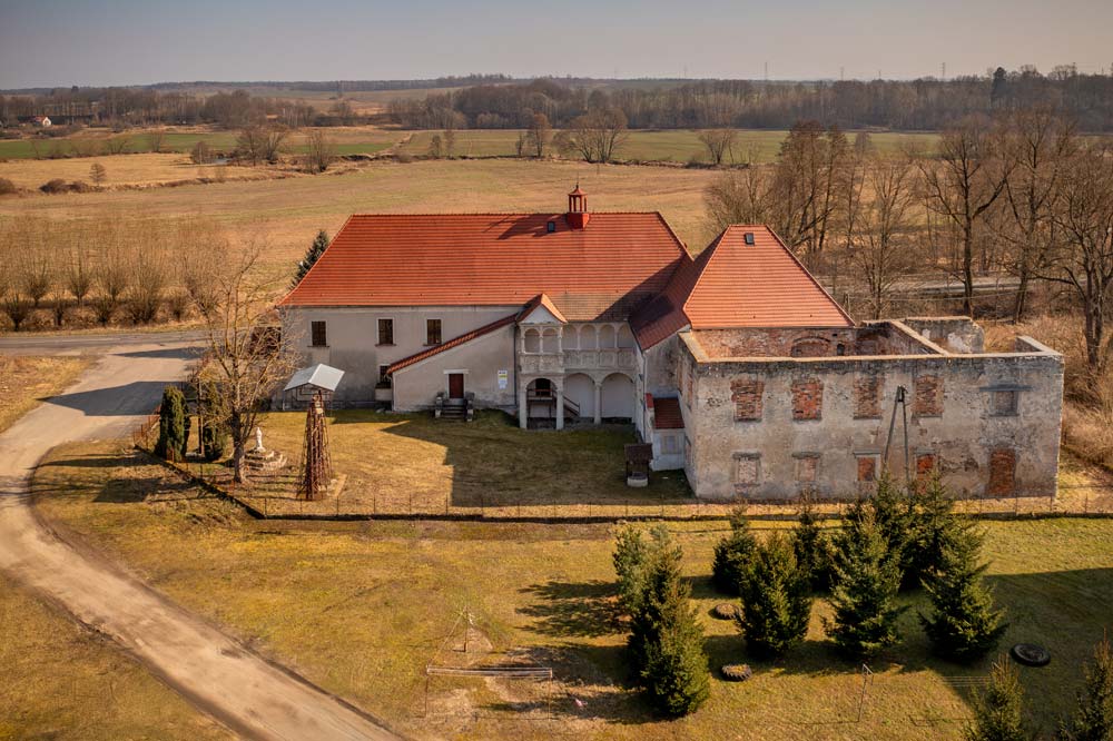 Renesansowy dwór w Nawojowie Łużyckim z lotu ptaka – skrzydło zaadaptowane na kościół z czerwonym dachem, obok zrujnowane skrzydło zachodnie bez dachu, wśród pól Górnych Łużyc