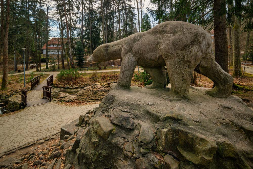 Rzeźba niedźwiedzia polarnego na kamiennym kopcu w Parku Leśnym w Polanicy-Zdroju, w tle drewniany mostek