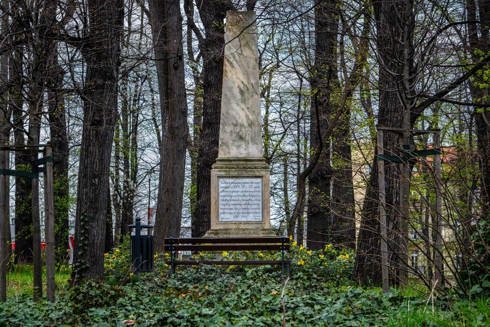 Piaskowcowy obelisk w Parku Młodzieżowym w Świdnicy widziany między pniami starych drzew, przed nim ławka