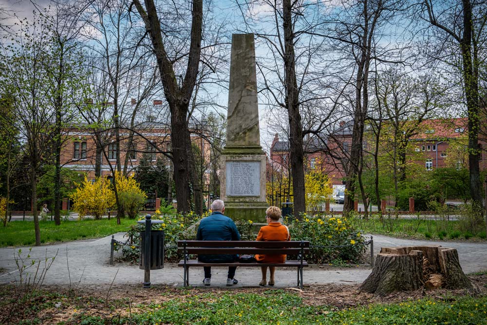 Para siedząca na ławce naprzeciw obelisku w Parku Młodzieżowym w Świdnicy, w tle zabudowa miejska
